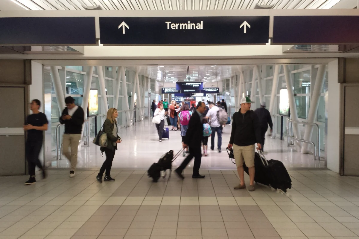 Travelers carrying and pulling luggage walk through a busy airport corridor beneath a sign directing them toward the terminal.
