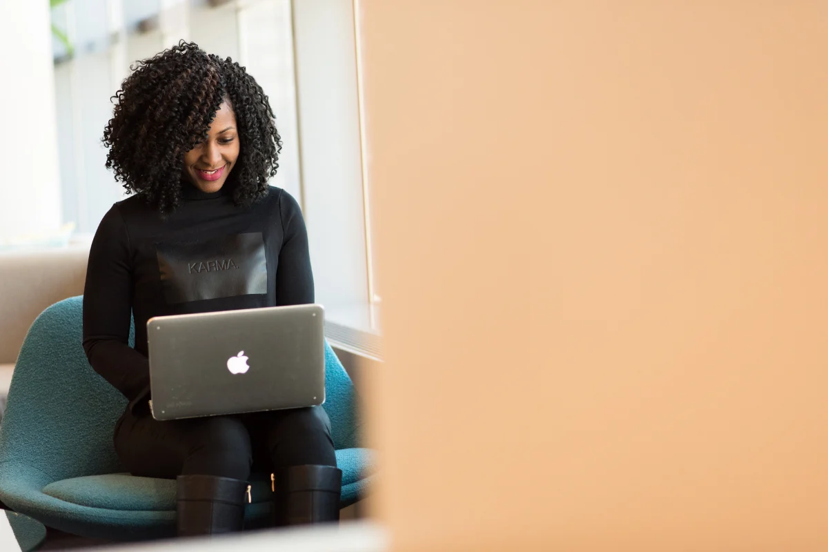 Woman smiles while working on a laptop from a lounge chair in a bright, modern indoor space.