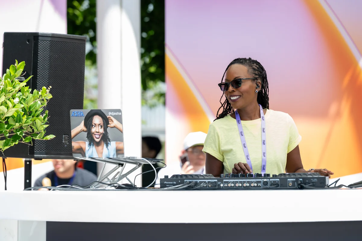 DJ smiles while mixing music at an outdoor event, standing behind audio equipment against a colorful backdrop.