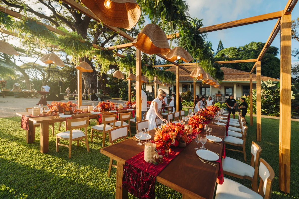 Elegant outdoor dining setup with long wooden tables, vibrant floral arrangements, hanging greenery, and woven pendant lights in a garden setting.