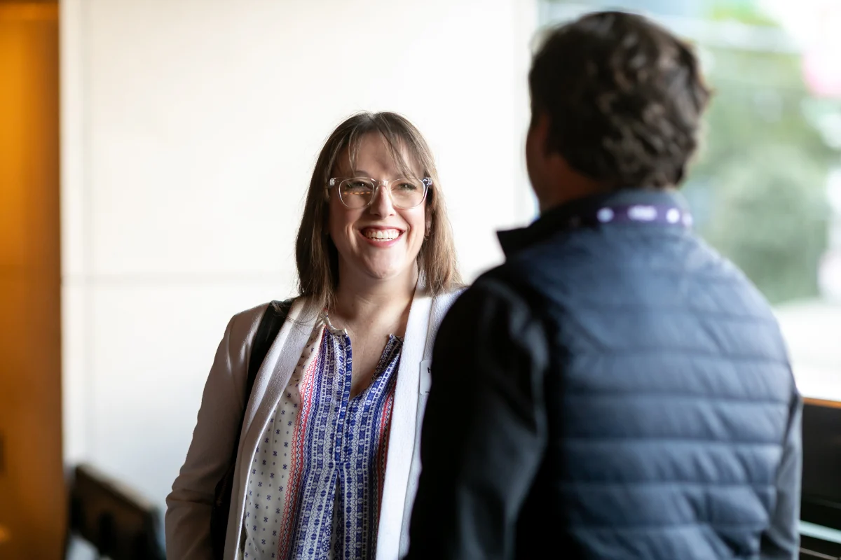 Two conference attendees have a friendly conversation, with one smiling as they speak in a bright indoor event space.