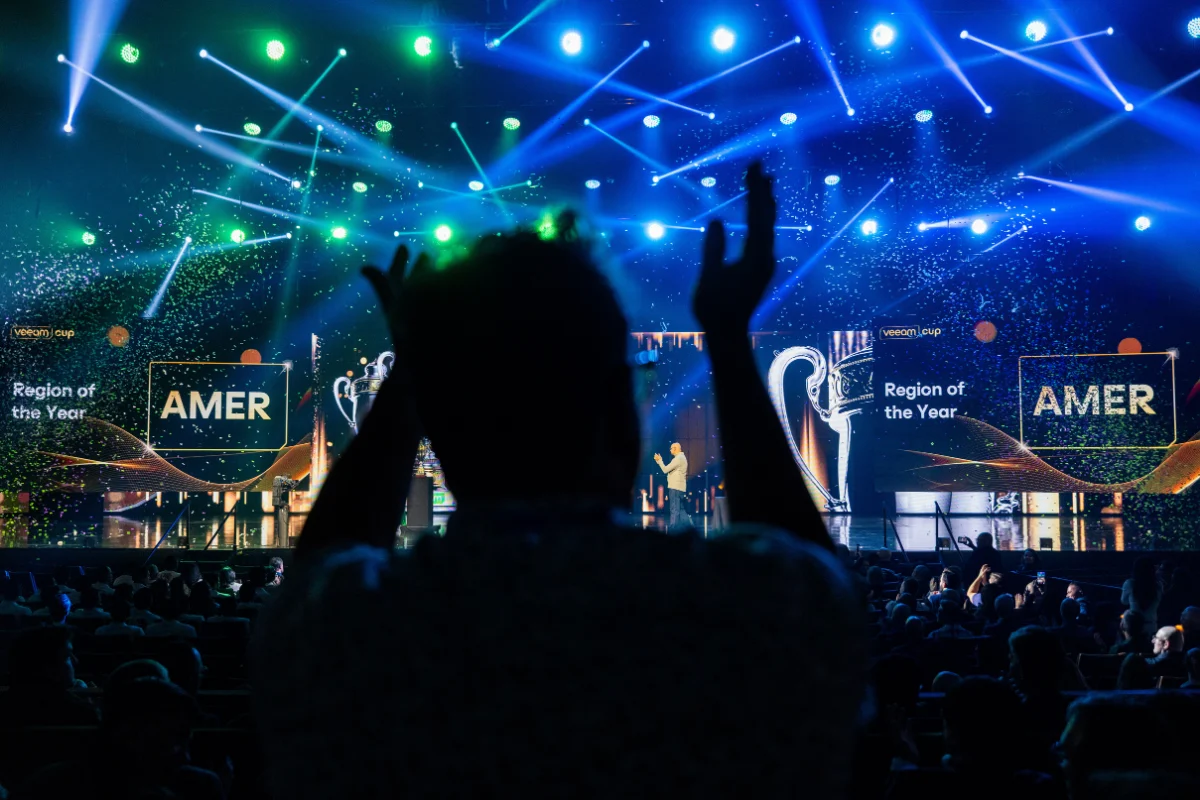 Silhouetted attendee applauds in front of a brightly lit event stage displaying “AMER Region of the Year,” with blue and green lighting effects and a seated audience.