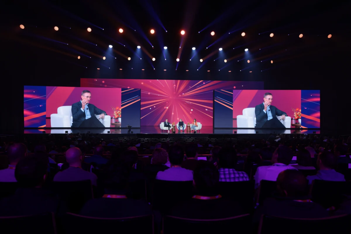 Wide view of a conference audience facing a large stage with vibrant pink and purple screens displaying a panel discussion.