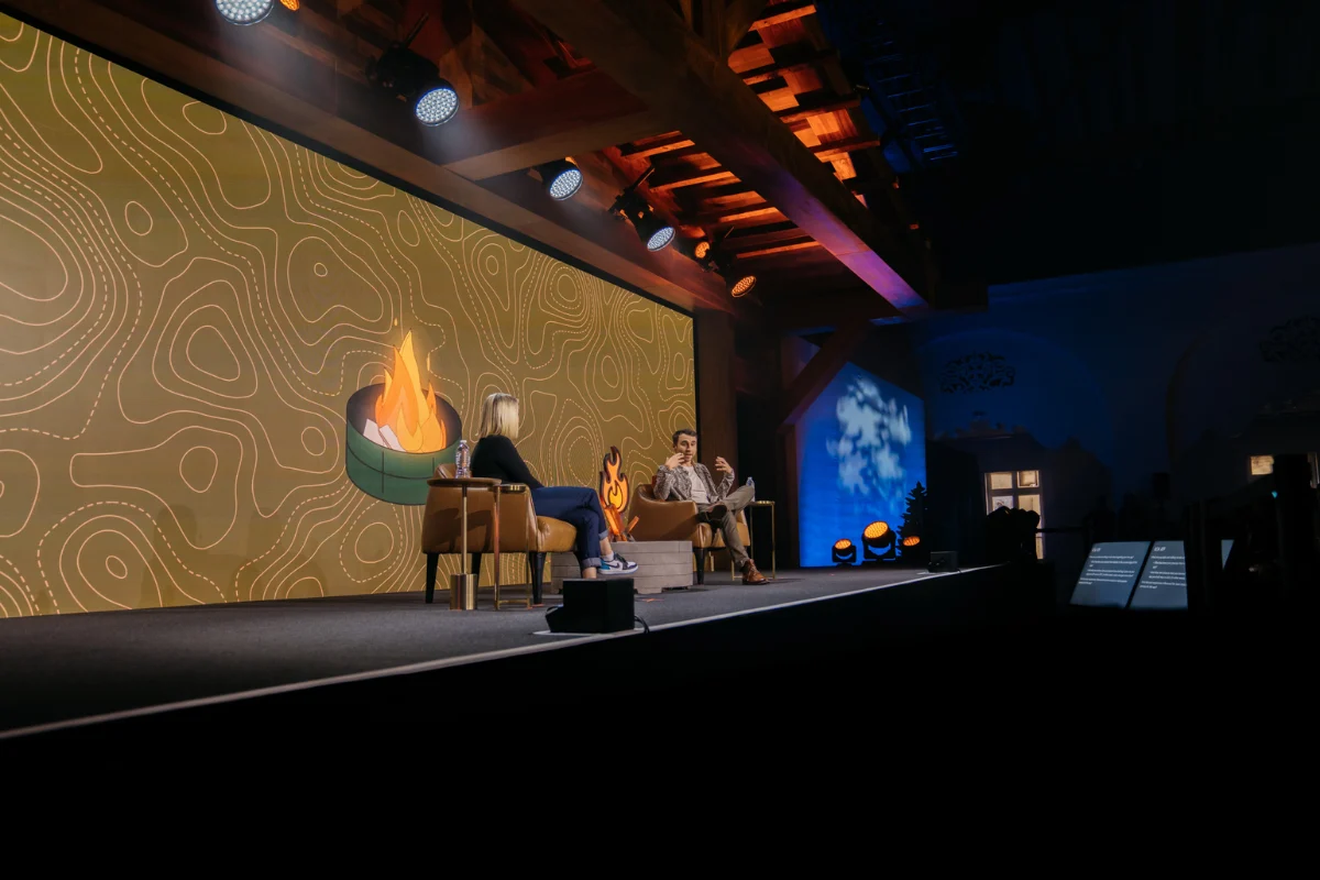 Two speakers sit onstage during a fireside chat, framed by warm lighting and a large illustrated campfire backdrop.