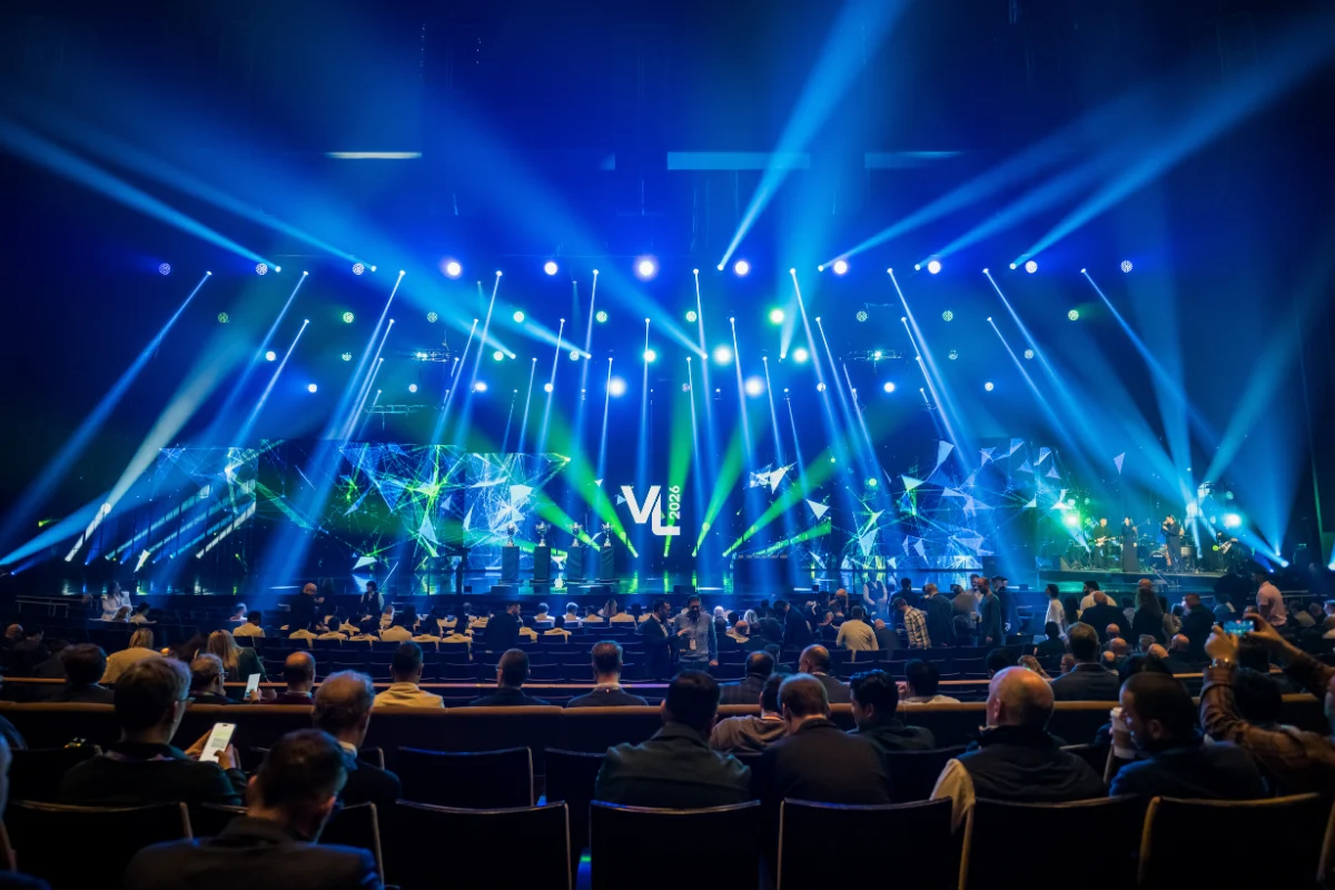 Large conference audience faces an expansive stage illuminated with dramatic blue and green lighting and abstract digital graphics.
