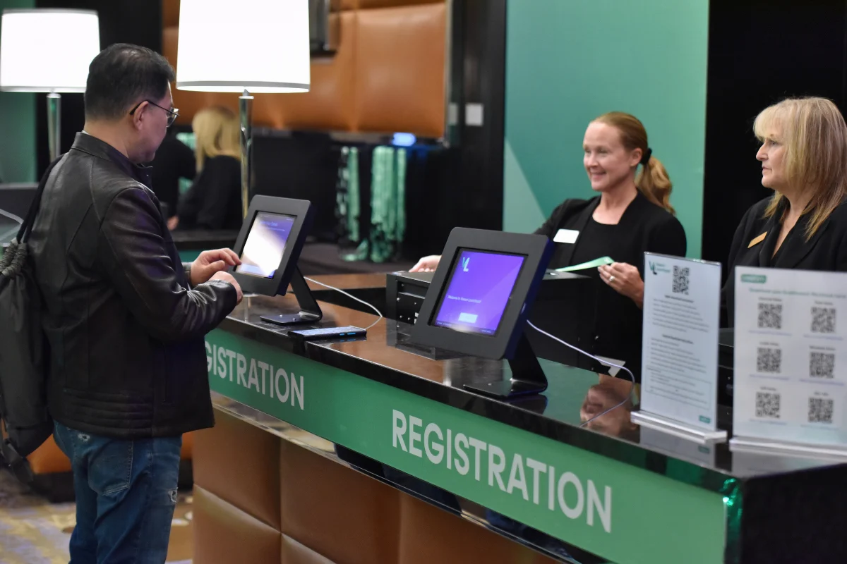 Attendee checks in at a conference registration desk with digital kiosks while event staff assist behind the counter.