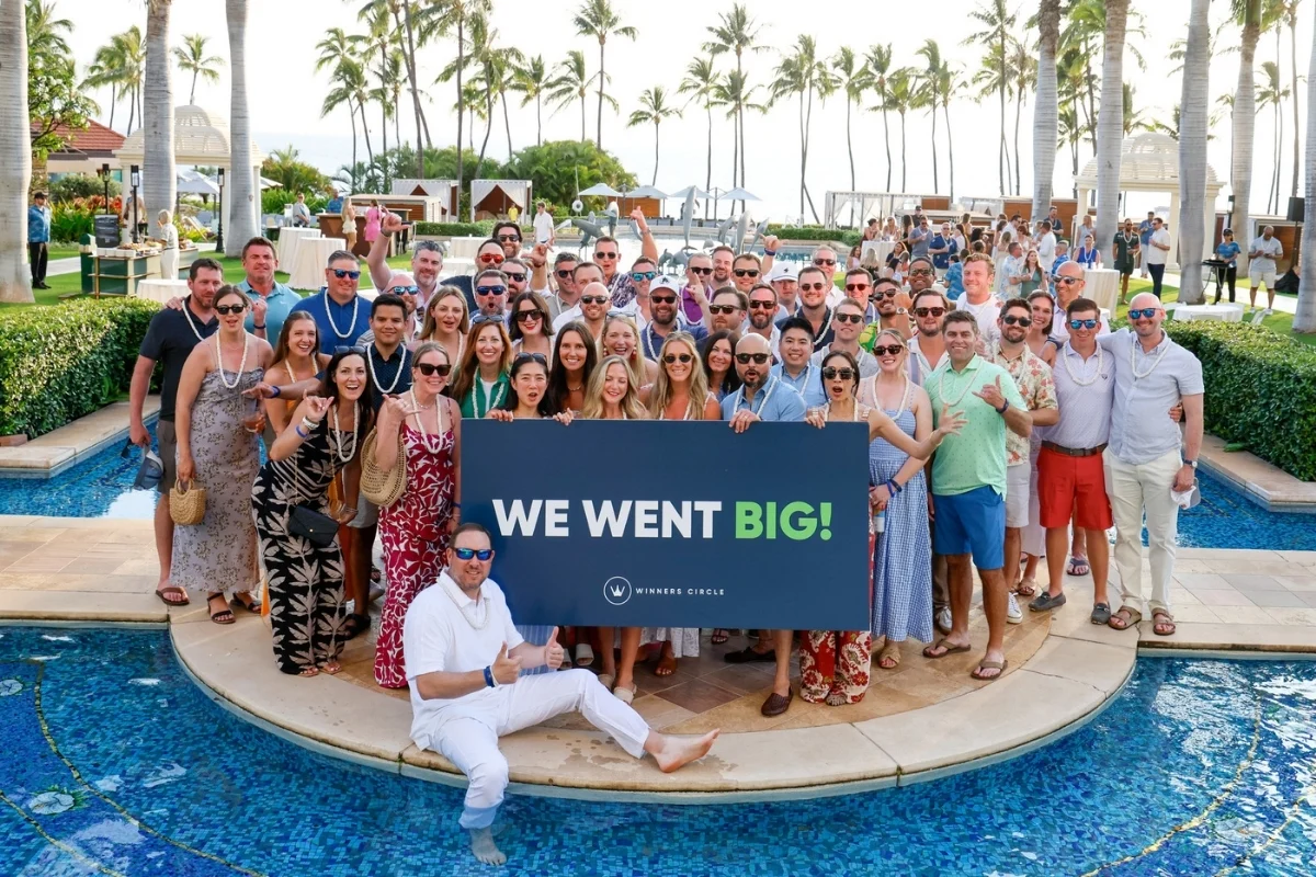 Large group of attendees pose together by a resort pool holding a “We Went Big!” Winners Circle sign, with palm trees and the ocean in the background.