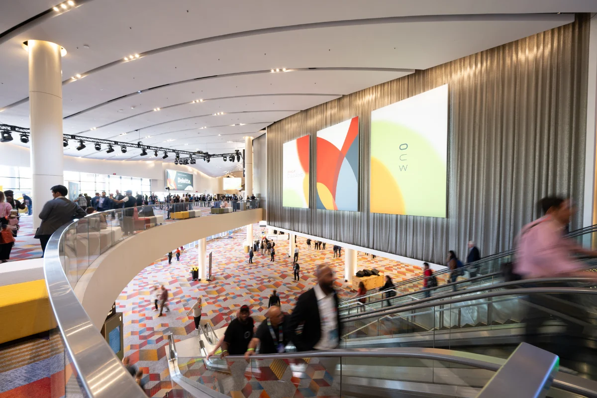 Attendees move through a bright, multi-level convention center with escalators, colorful carpeting, and large event graphics.