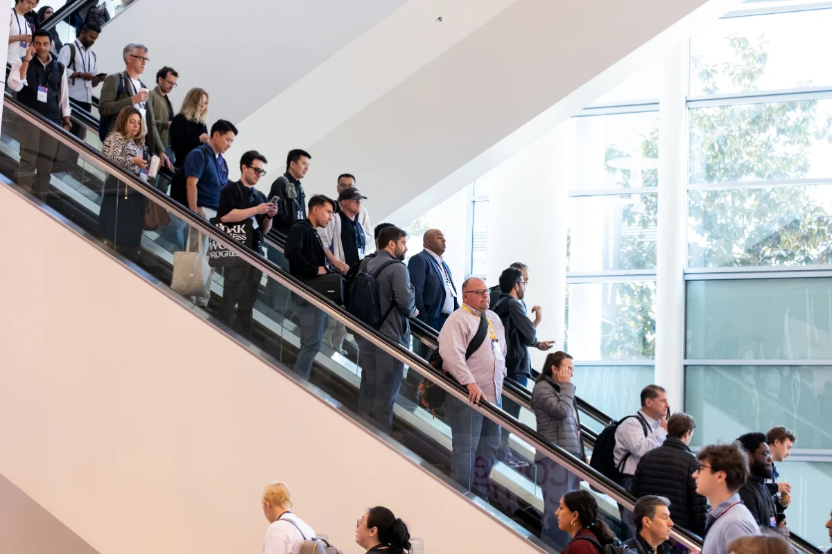 Conference attendees ride a crowded escalator through a bright, modern convention center.