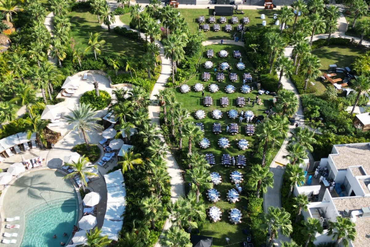 Aerial view of an outdoor event setup with dining tables and seating arranged on a lush resort lawn surrounded by palm trees and a pool.