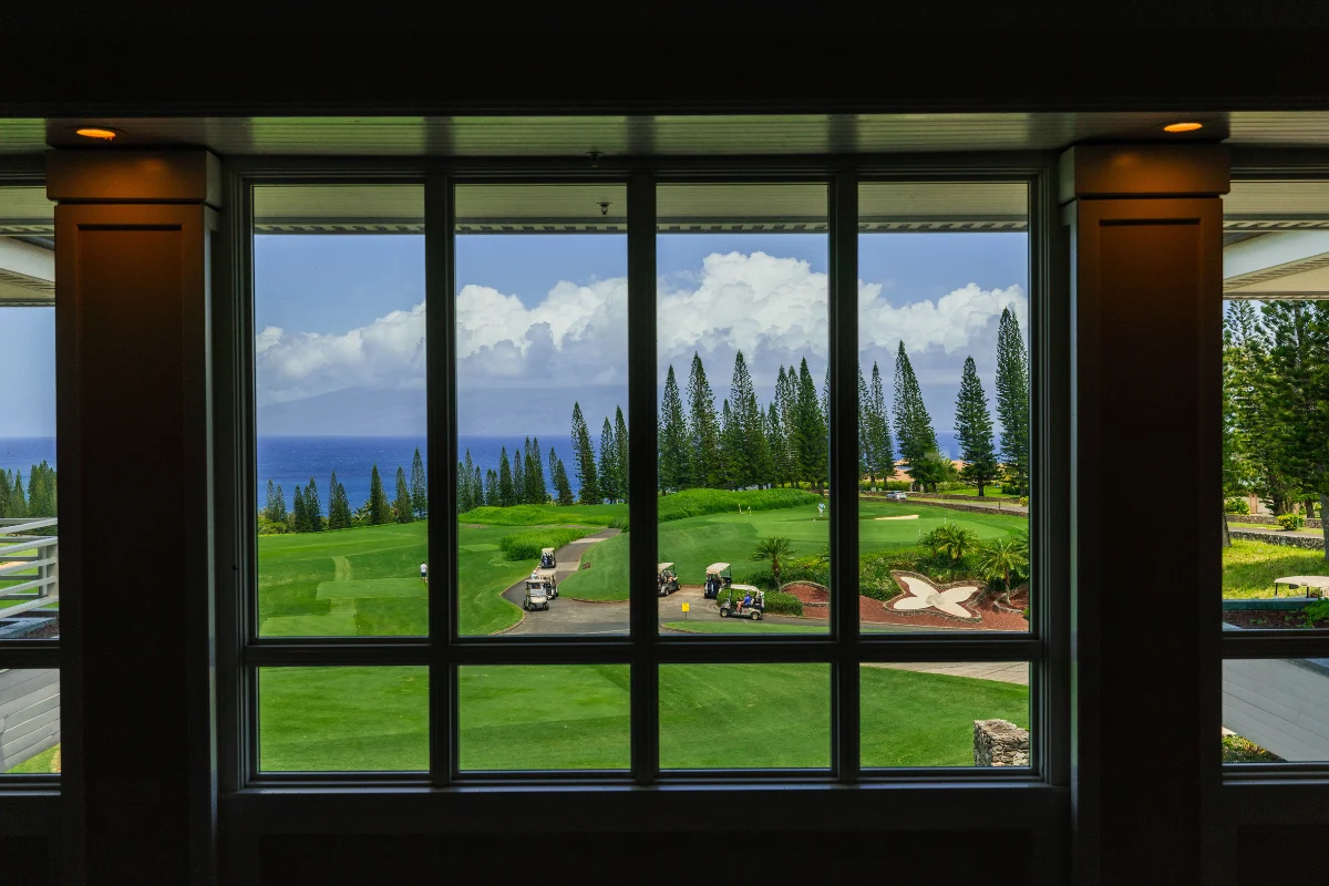 View through large windows overlooking a lush golf course with golf carts, pine trees, and the ocean in the distance.